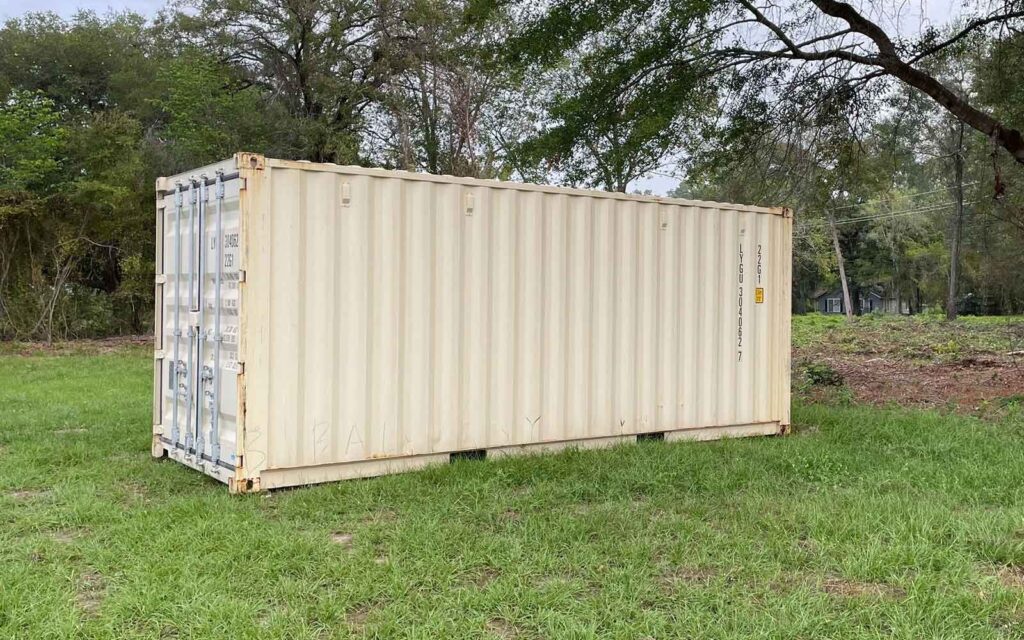 Beige shipping container on a grassy field with trees and a house visible in the distance in a rural setting.