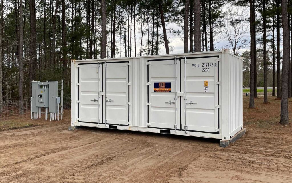 White shipping container on a dirt lot beside a small electrical panel, surrounded by tall pine trees in a wooded area.