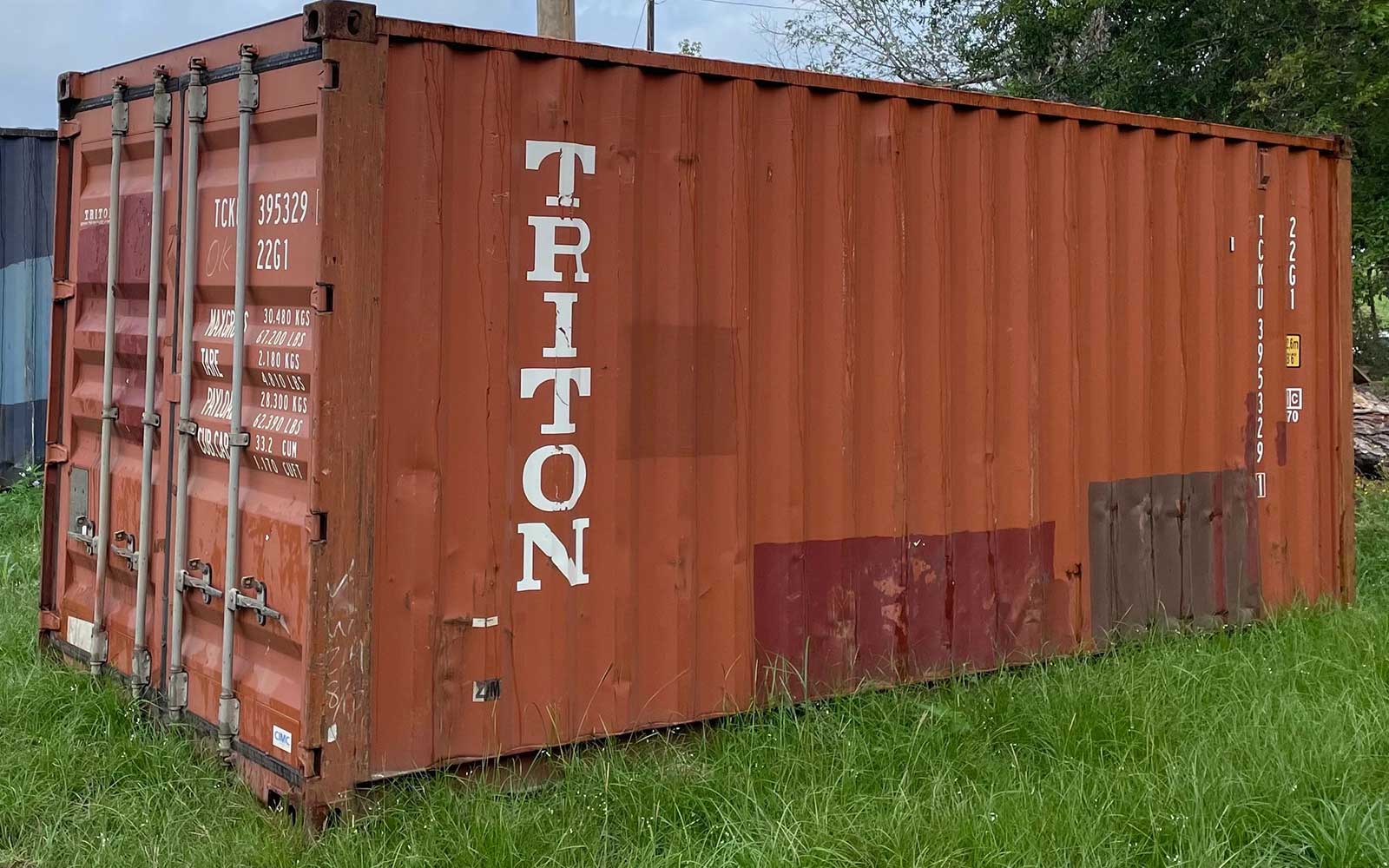 Rust-colored shipping container on a grassy field with white vertical lettering along the side.