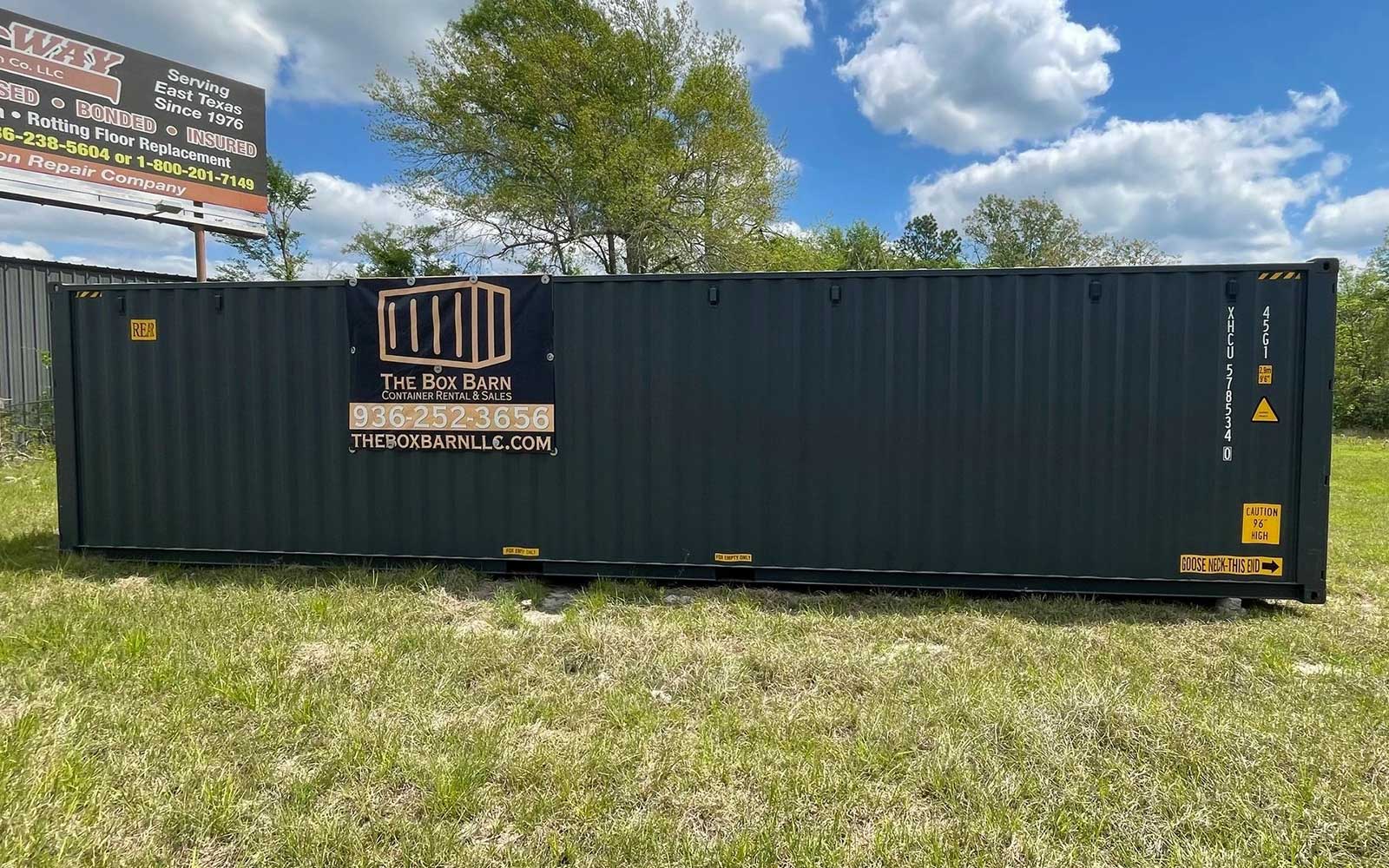 Long dark blue shipping container on a grassy lot with a banner reading The Box Barn LLC and a phone number