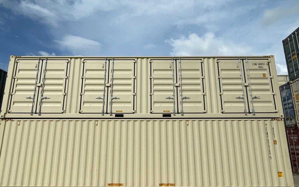Two beige shipping containers stacked on top of another container, with multiple metal doors and locking bars under a blue sky habitatized by light clouds.