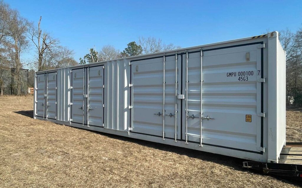Long white shipping container on a grassy field with multiple doors and a clear blue sky behind it.