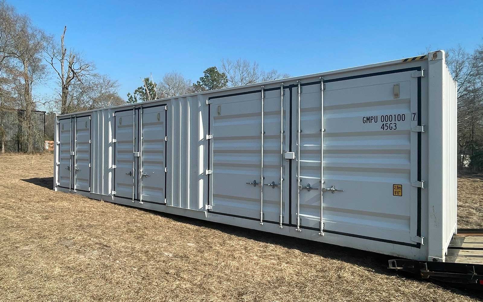 Long white shipping container on a grassy field with multiple doors and a clear blue sky behind it.