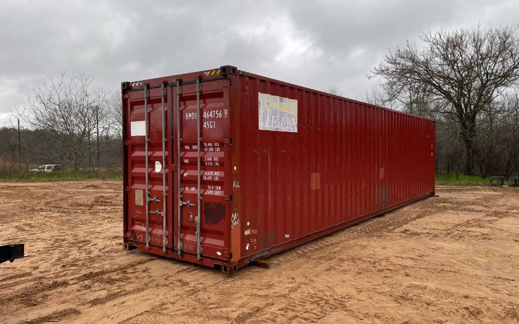 Red shipping container on a dirt lot under a cloudy sky, with bare trees in the background.